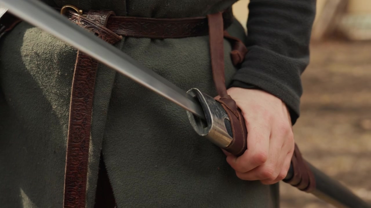 Una vista all'altezza della vita mostra una persona che stabilizza il fodero di una spada con una mano, con le dita avvolte attorno all'elsa accanto al fodero in pelle, evidenziando la texture del costume, il metallo usurato e il movimento di preparazione alla luce naturale.