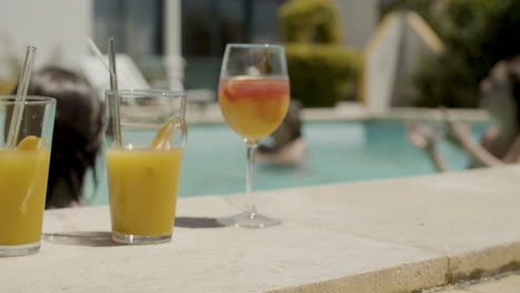 close up of orange juices and cocktails on poolside while in the background a happy family playing with ball in swimming pool