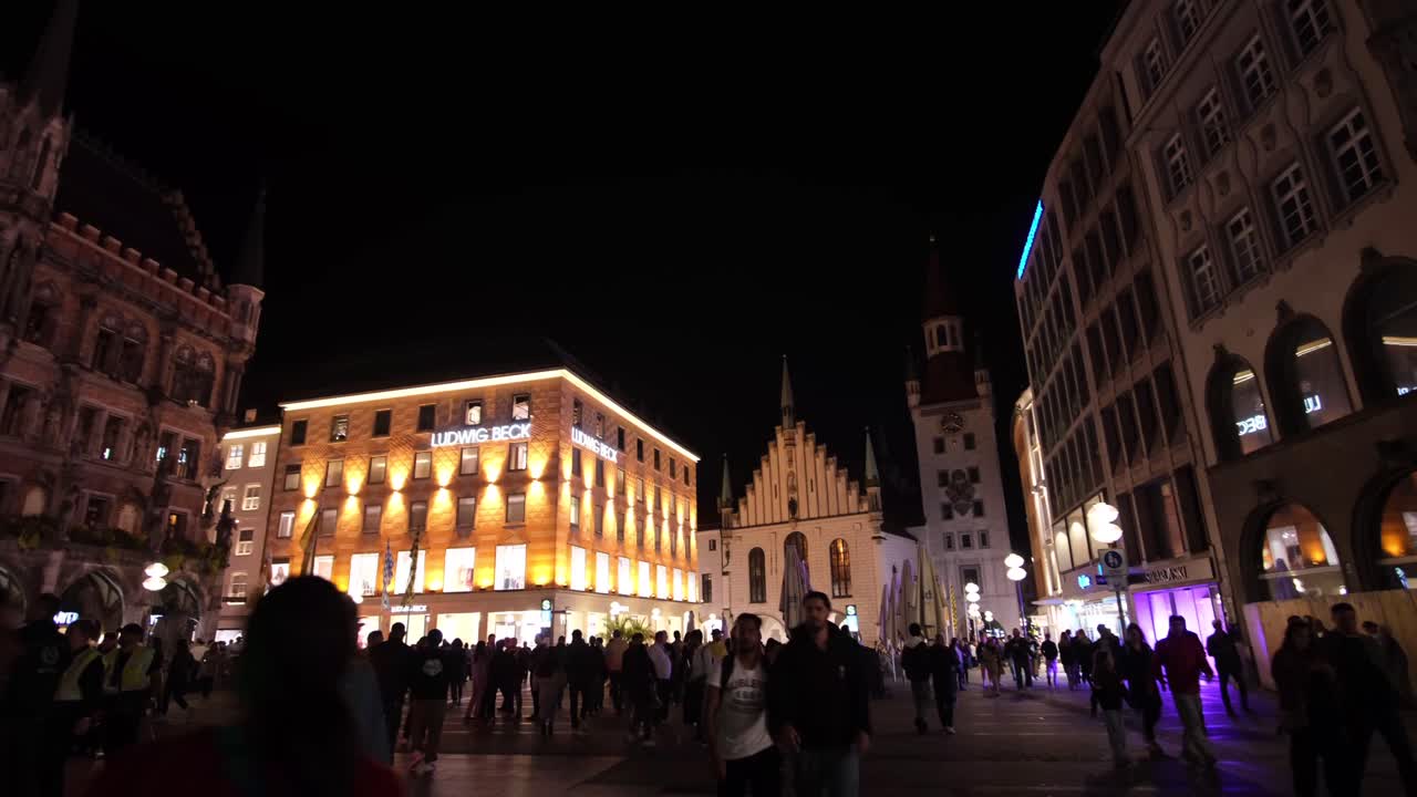 A night scene with people strolling past Ludwig Beck’s light-yellow building, framed by towering Bavarian structures on both sides.