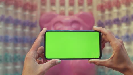 hands holding a smartphone with a green screen in front of a blurred pink bear sculpture