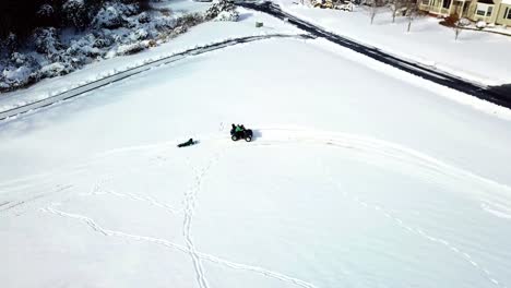 a drone, aerial, birds eye view tracking an atv 4 wheeler with a child riding on the back and pulling another child riding on their belly on a winter sled on the snow covered ground in the country