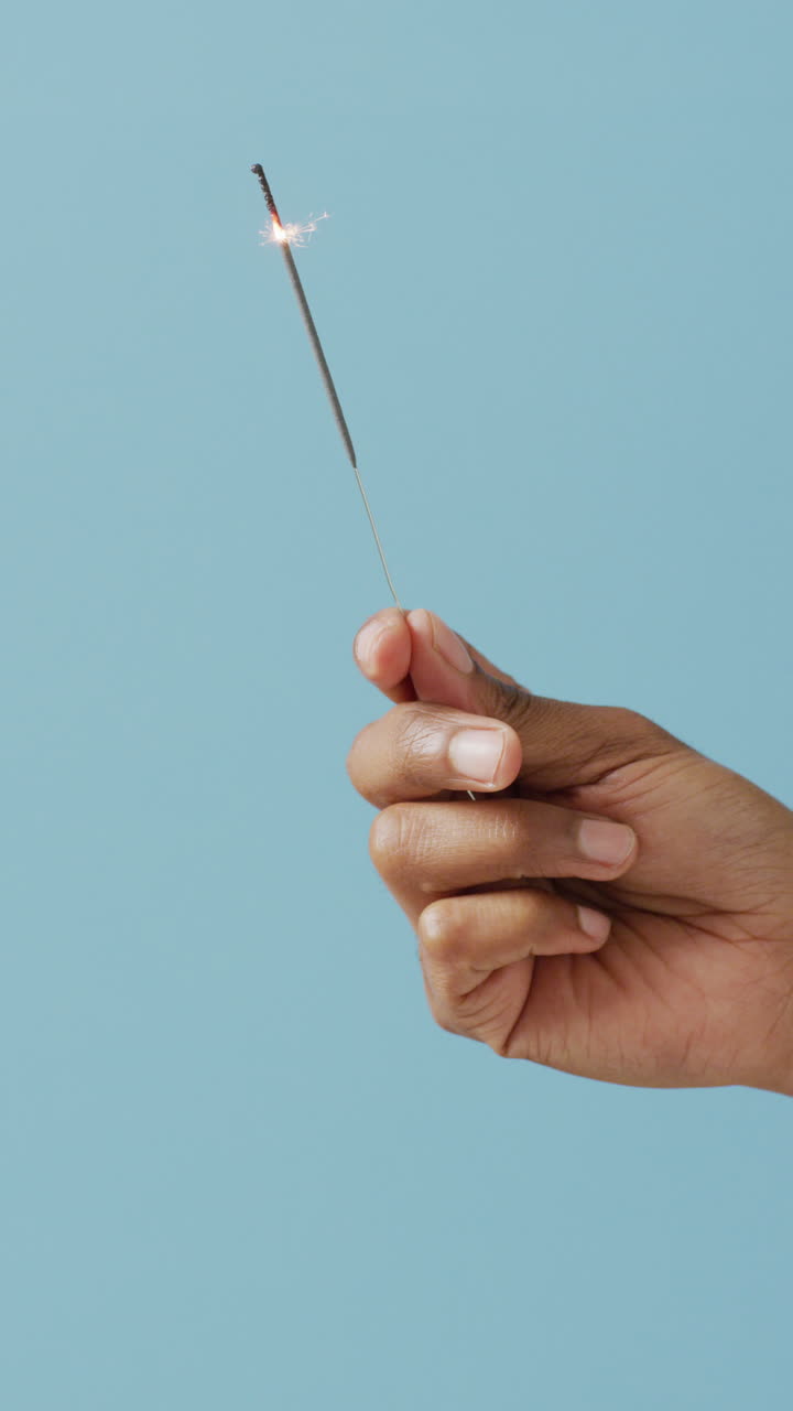 video vertical de la mano de una persona afroamericana sosteniendo una chispa encendida sobre un fondo azul
