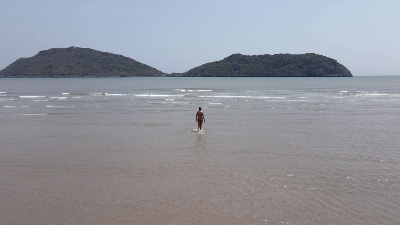 Lone woman in swim suit walks into ocean at sandy tropical beach