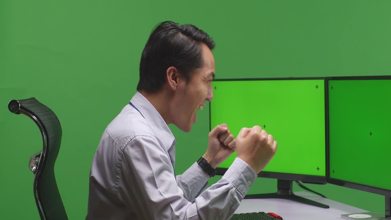 primer plano de un hombre asiático feliz celebrando y bailando mientras trabaja con un simulacro de varios monitores de computadora en un estudio de pantalla verde