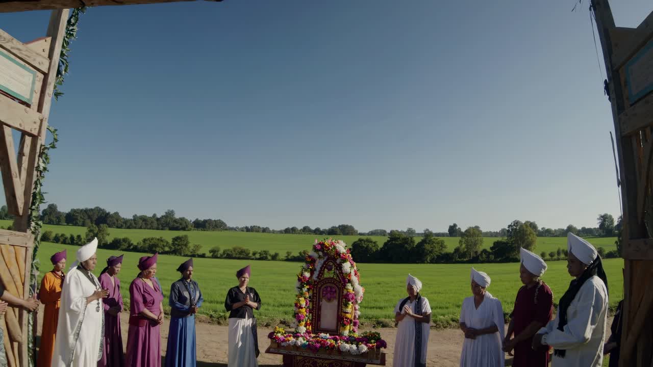 Group of people dressed in vibrant traditional clothing celebrating a religious ceremony in a lush green field under a bright blue sky, joyfully carrying a beautifully adorned floral structure