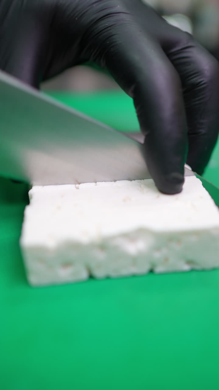 Vertical footage Professional chef's hands wearing protective gloves slicing feta cheese on green cutting board, showcasing precise food preparation skills