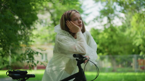 young woman in translucent raincoat rests her head on her hand, leaning thoughtfully on her bicycle handlebar, lost in contemplation, background features lush green trees