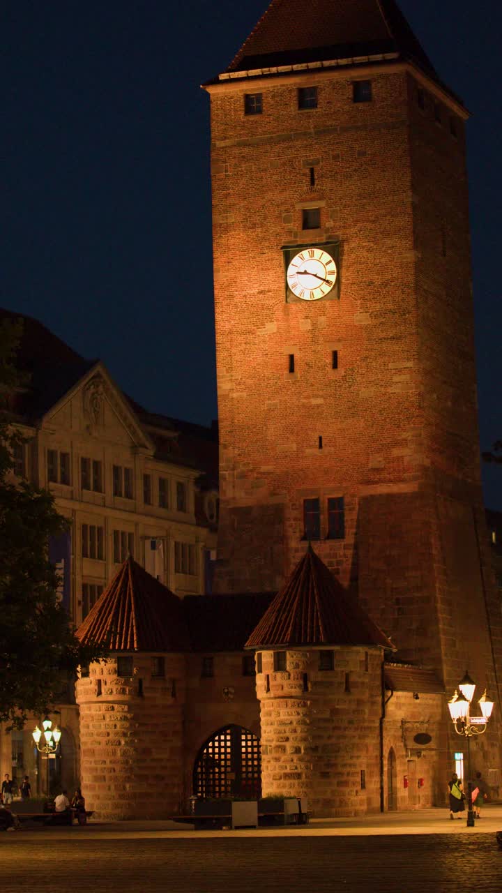 Illuminated clock tower and old town square at night, warm lighting, slow camera pan right