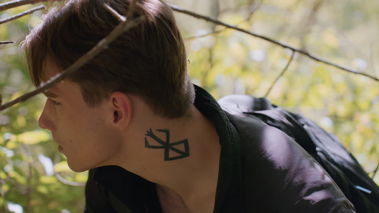 Young soldier with tattoo on neck bends cautiously under tree branch in sunlit forest, scanning surroundings attentively while partially hidden in foliage, conveying alertness and strategic movement