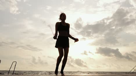 Unrecognizable-athletic-woman-working-out-on-the-jump-rope-against-the-son-by-the-beach-in-Slow-Motion.-Girl-jumping-on-a