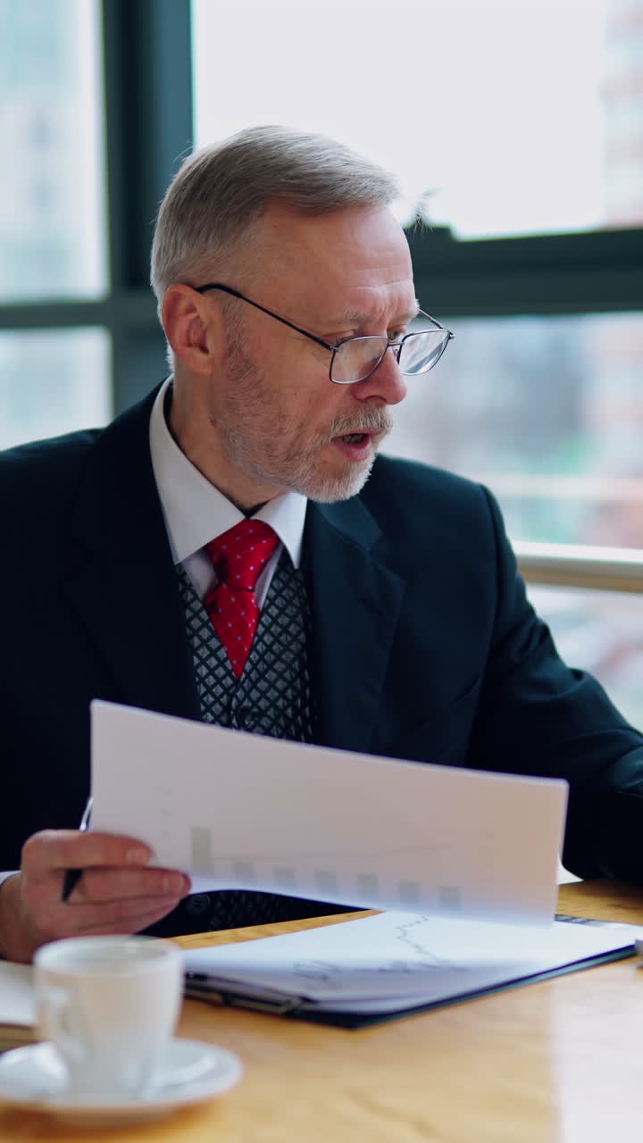 Businessman is checking a printed report or document while sitting at table, working on paper work at a laptop explaining his point of view. Video in an office. Vertical video