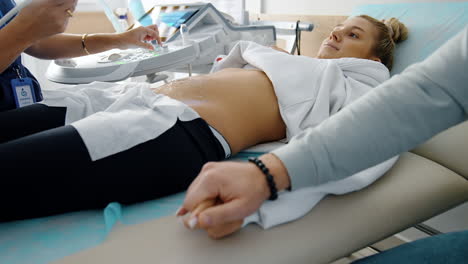 beautiful pregnant lady undergoing ultrasound examination. male hand takes his wife's hand showing love and support.
