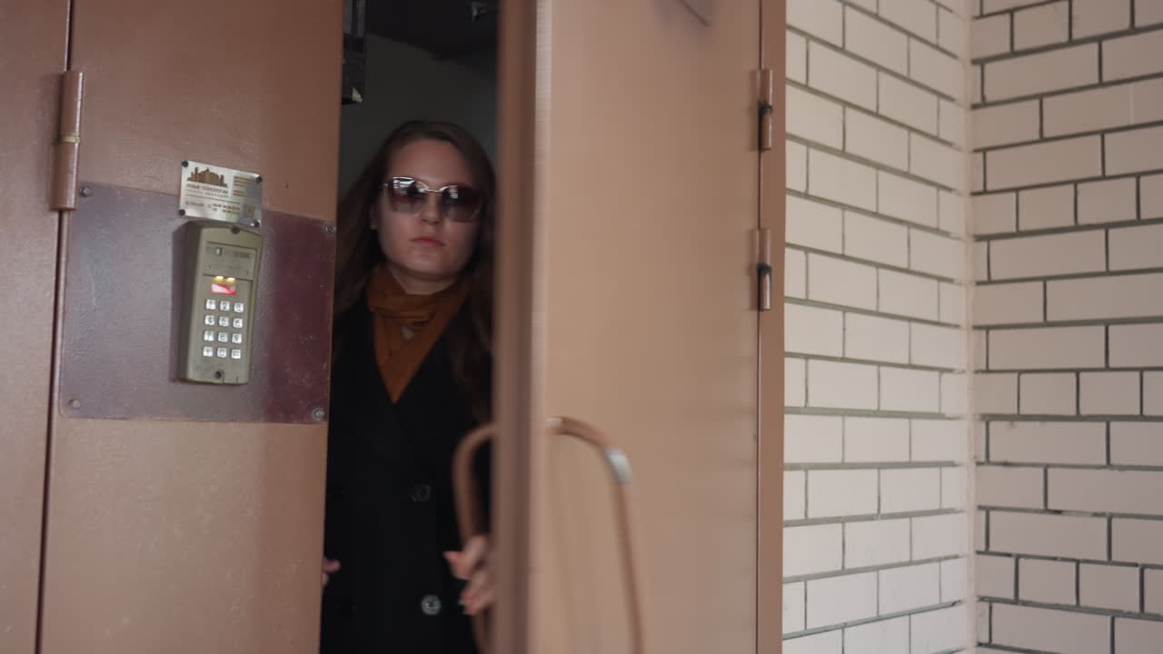 En una entrada de seguridad, se muestra un intercomunicador con teclado y dígitos rojos mientras una empresaria cruza una puerta hacia la oficina. Se ven la manija y las bisagras junto a la pared de ladrillo, en una escena de control de acceso que sugiere la introducción de un código.