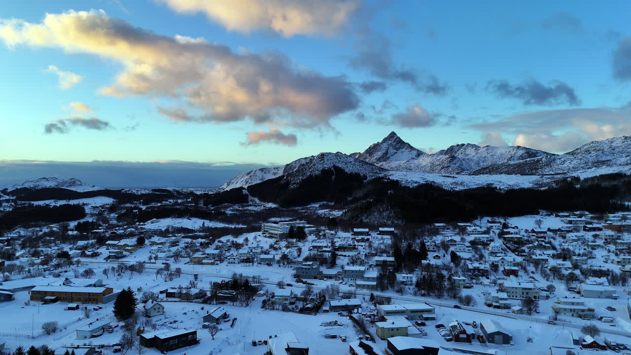 Cinematic aerial view of winter town in Lofoten, Norway, surrounded by snow-covered houses, mountains, and dramatic arctic landscape under soft evening light