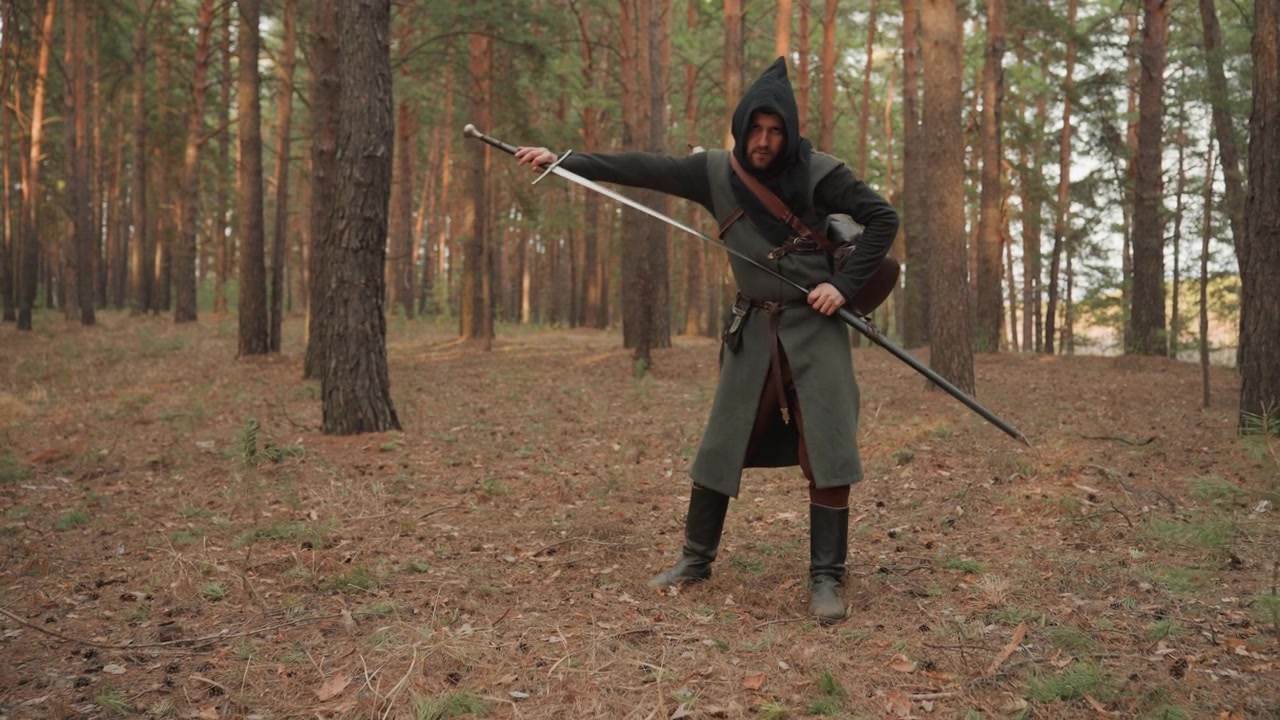 Figura encapuzada em manto estilo medieval move-se por um bosque ralo de pinheiros, manuseando uma besta enquanto passa de um passo cauteloso para uma postura ereta de prontidão com foco alerta, como um arqueiro encapuzado