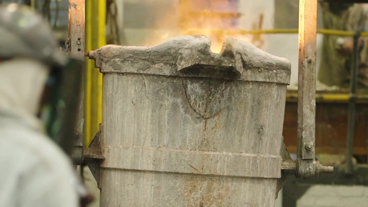 Large metal casting ladle sits upright with orange flames escaping from the top in an active industrial workshop.