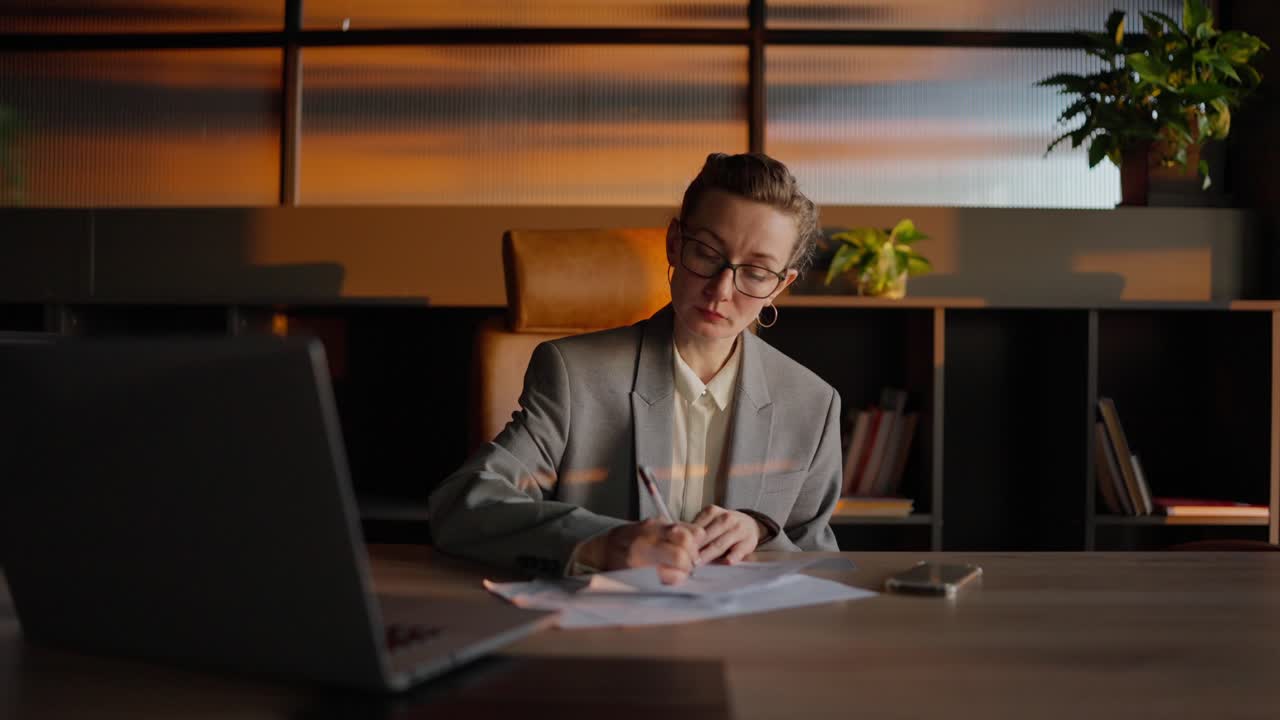 Confident middle-aged business woman with glasses in a gray jacket and business uniform sits at a wooden table and writes down her ideas in front of a laptop in a sunny office