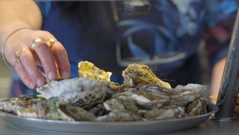 Close-up-shot-of-a-plate-of-oysters-where-a-person-with-a-hand-adorned-with-gold-rings-adding-more-bits