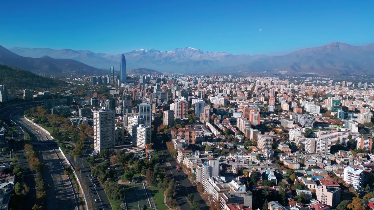 Volo in avanti su una vasta espansione urbana residenziale con il corridoio autostradale Alameda e la torre Costanera Center che si erge contro le Ande innevate. Santiago, Cile.