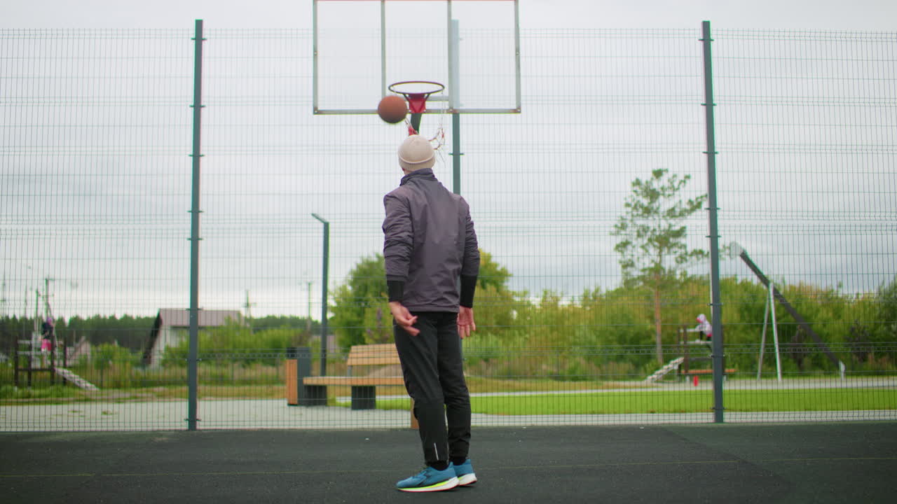 uomo che si allena a basket su un campo all'aperto con giacca e scarpe da ginnastica, cielo nuvoloso, parco recintato, che salta per raggiungere il canestro, rimbalzo della palla, espressione concentrata, routine di allenamento casuale, vegetazione urbana