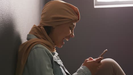 Asian-female-student-wearing-a-beige-hijab-leaning-against-a-wall-and-using-a-smartphone