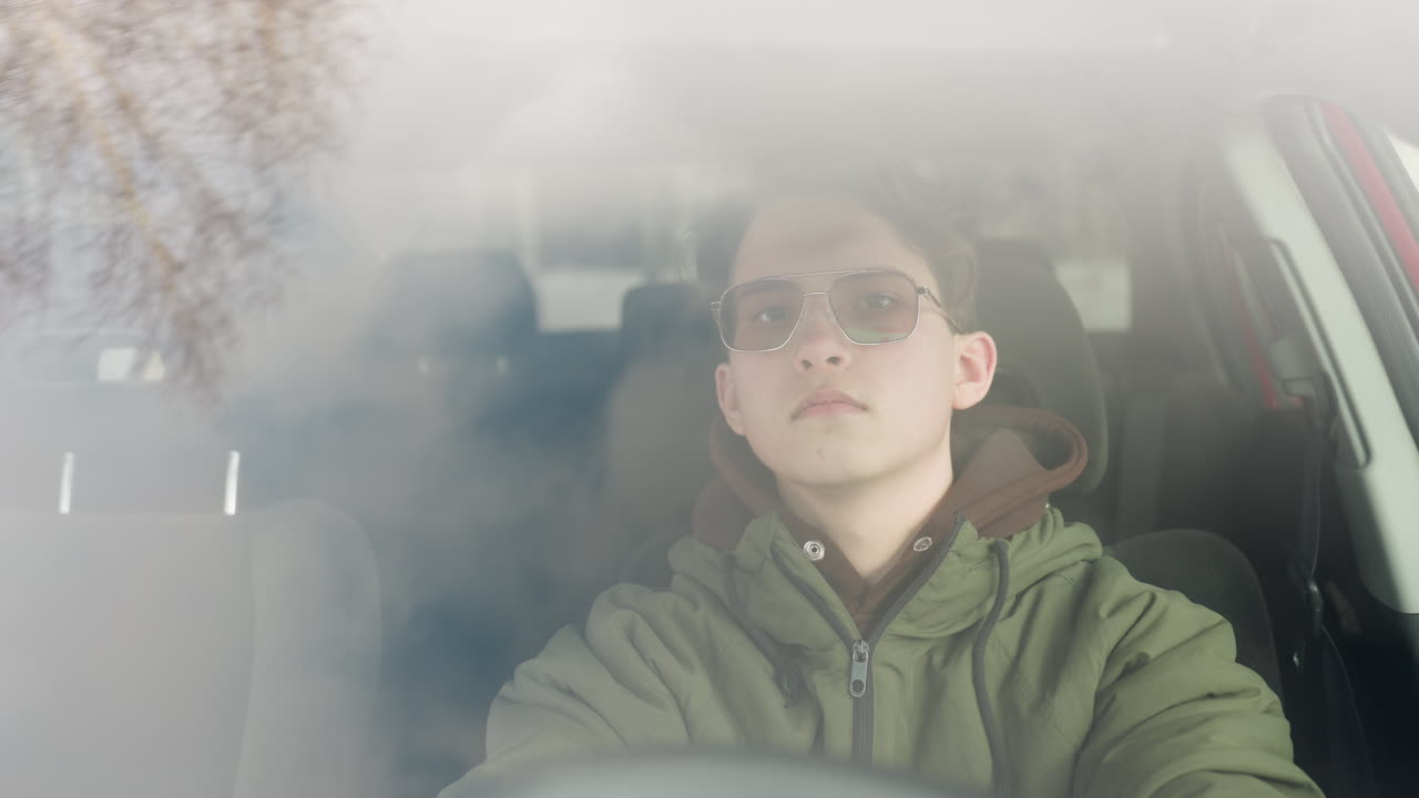 student seen through windshield sitting in driver seat with calm facial expression, wearing green winter jacket and glasses, natural reflection and diffused daylight visible on glass surface