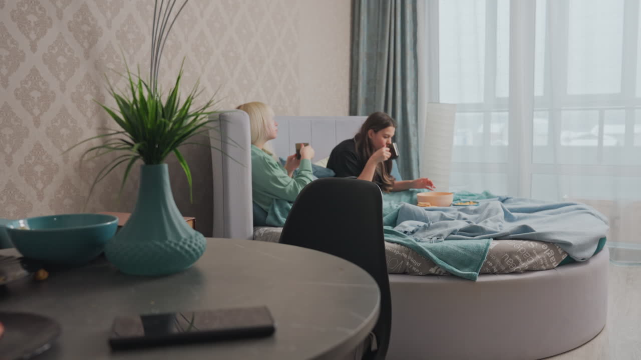 Two college girls enjoy lunch in bed wrapped in soft blankets while chatting, assorted snacks, bowls, and drinks are placed on bed, background features modern decor with natural daylight streaming in