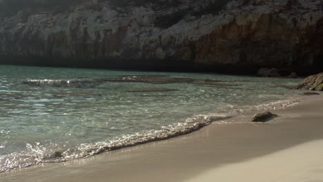 Sonniger-Blick-Auf-Den-Strand-Von-Caló-Des-Moro,-Der-Vom-Mittelmeer-Umspült-Wird,-Mit-Einigen-Reflexionen-Im-Wasser-Und-Felsformationen-Im-Hintergrund-Auf-Mallorca,-Balearen