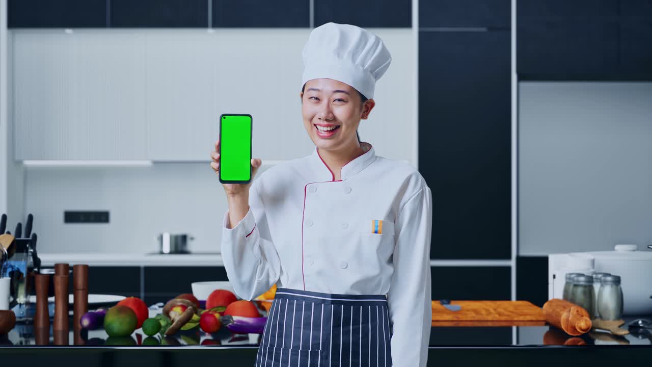 아시아 요리사 여성이 미소 짓고 가정 주방의 카메라에 녹색 화면 스마트폰을 보여줍니다.