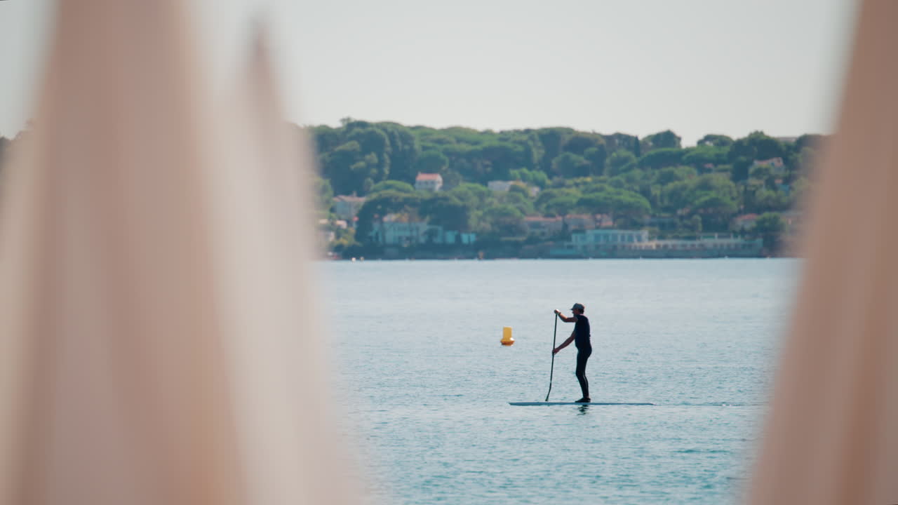 A man stand-up paddleboarding on tranquil blue water near a coastal town, framed by blurred umbrellas in the foreground