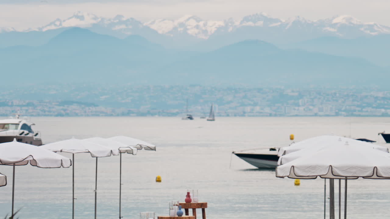 Close up of white sun umbrellas on the beach with multiple boats on the sea, the city and mountains on the background