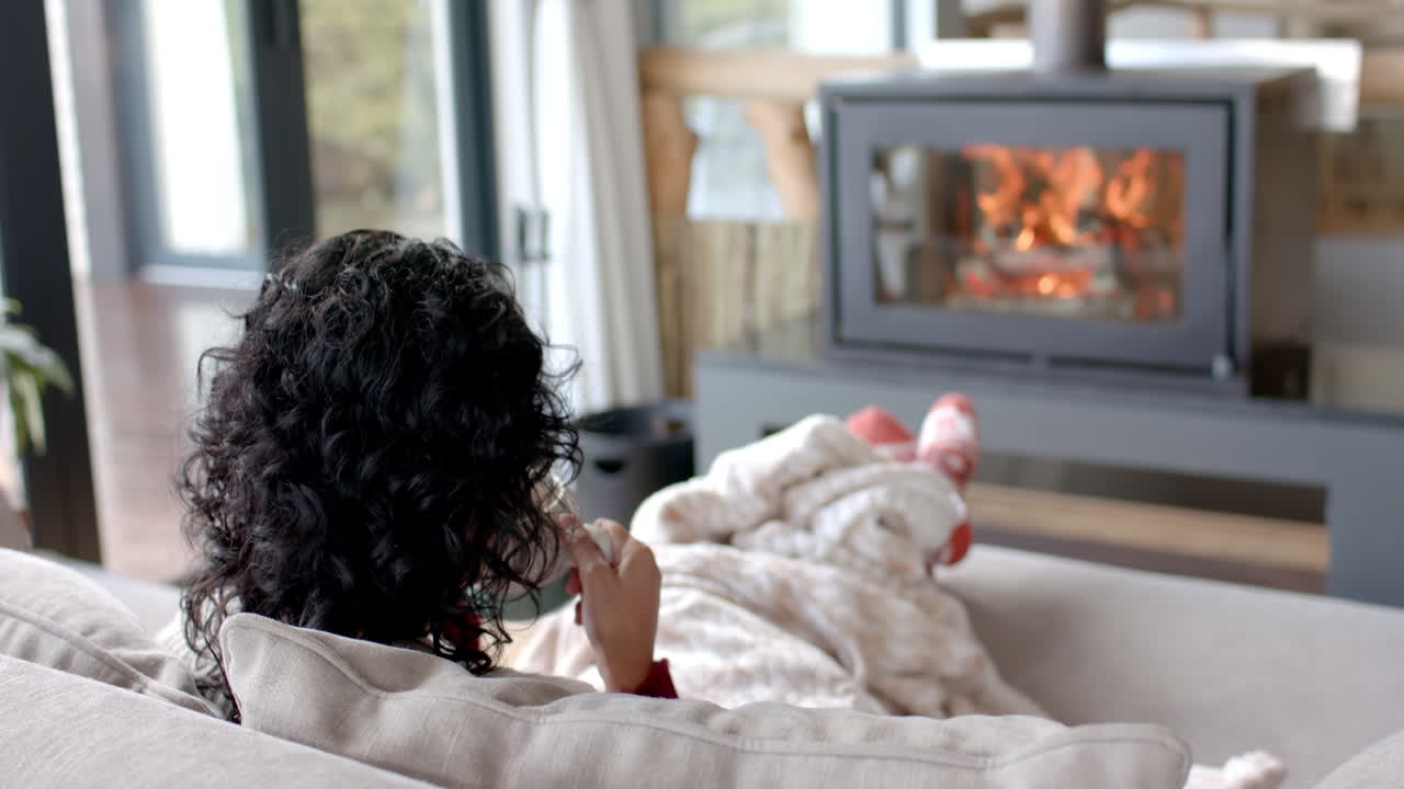 집에서 벽난로 앞에 담요 아래 소파에서 커피를 마시는 두 인종의 여성, 느린 동작