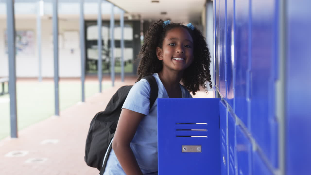 Biracial girl with curly hair stands by blue lockers in school, backpack on her shoulders