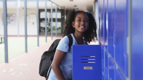 biracial girl with curly hair stands by blue lockers in school, backpack on her shoulders