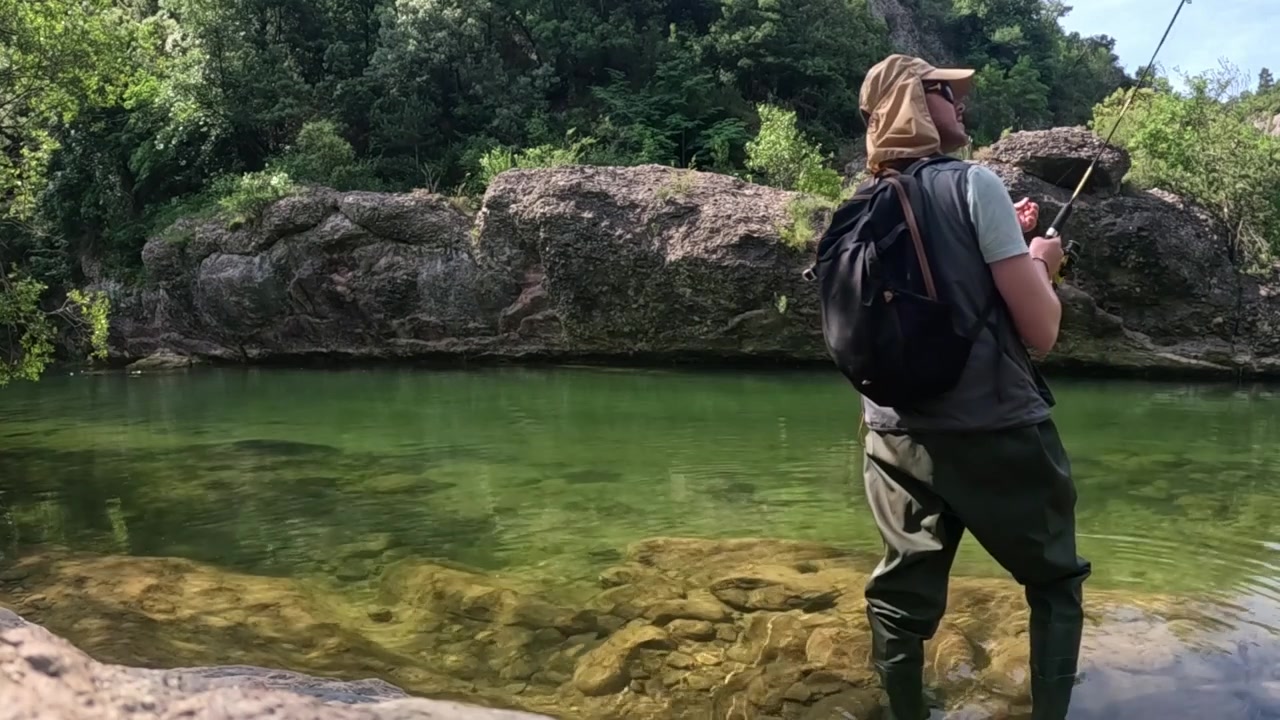 Static shot of an angler in waders raising a fishing rod from a clear river pool, with submerged stones and large boulders in view while targeting trout.
