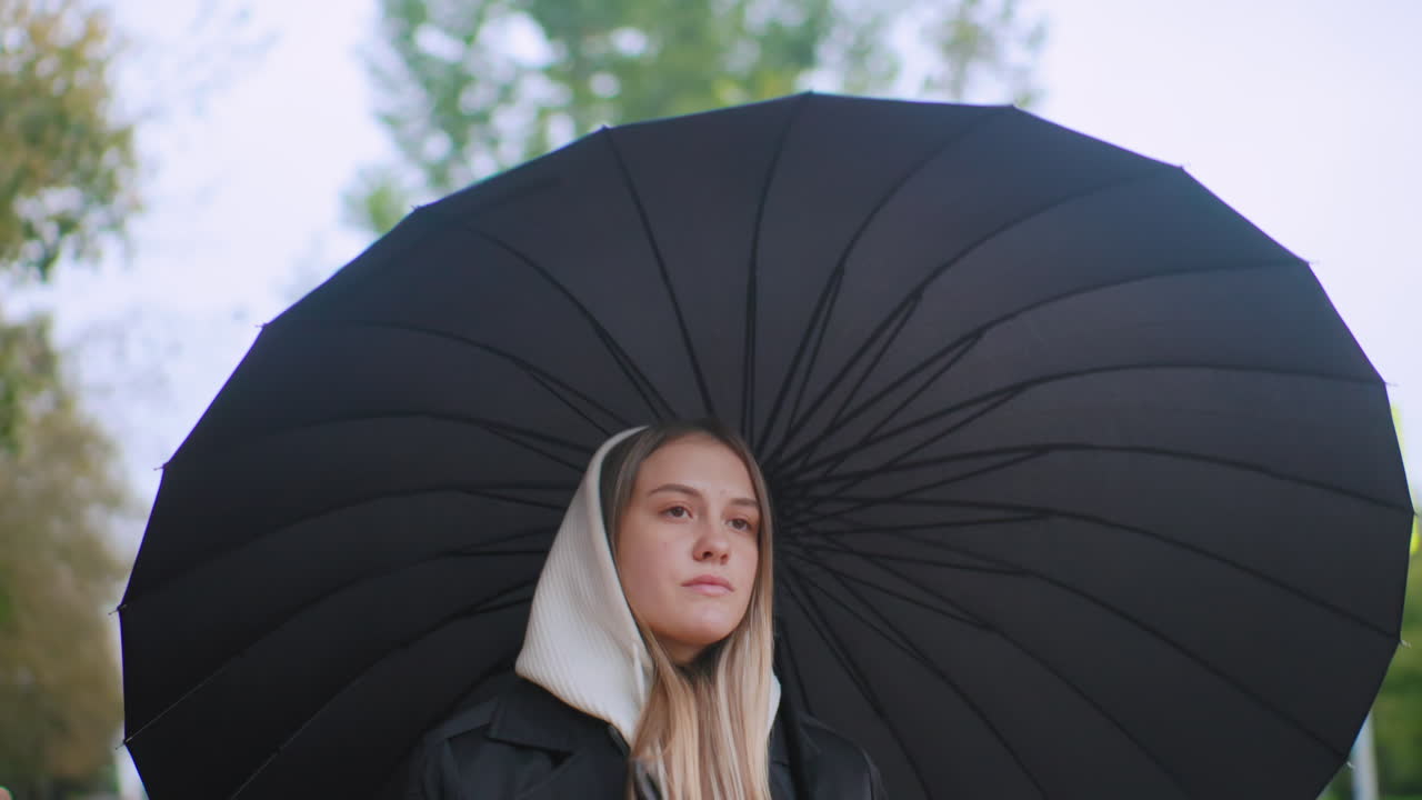 Young woman in white hood and black coat holds large black umbrella outdoors, looking calm and thoughtful against blurred green trees background, creating moody autumn atmosphere