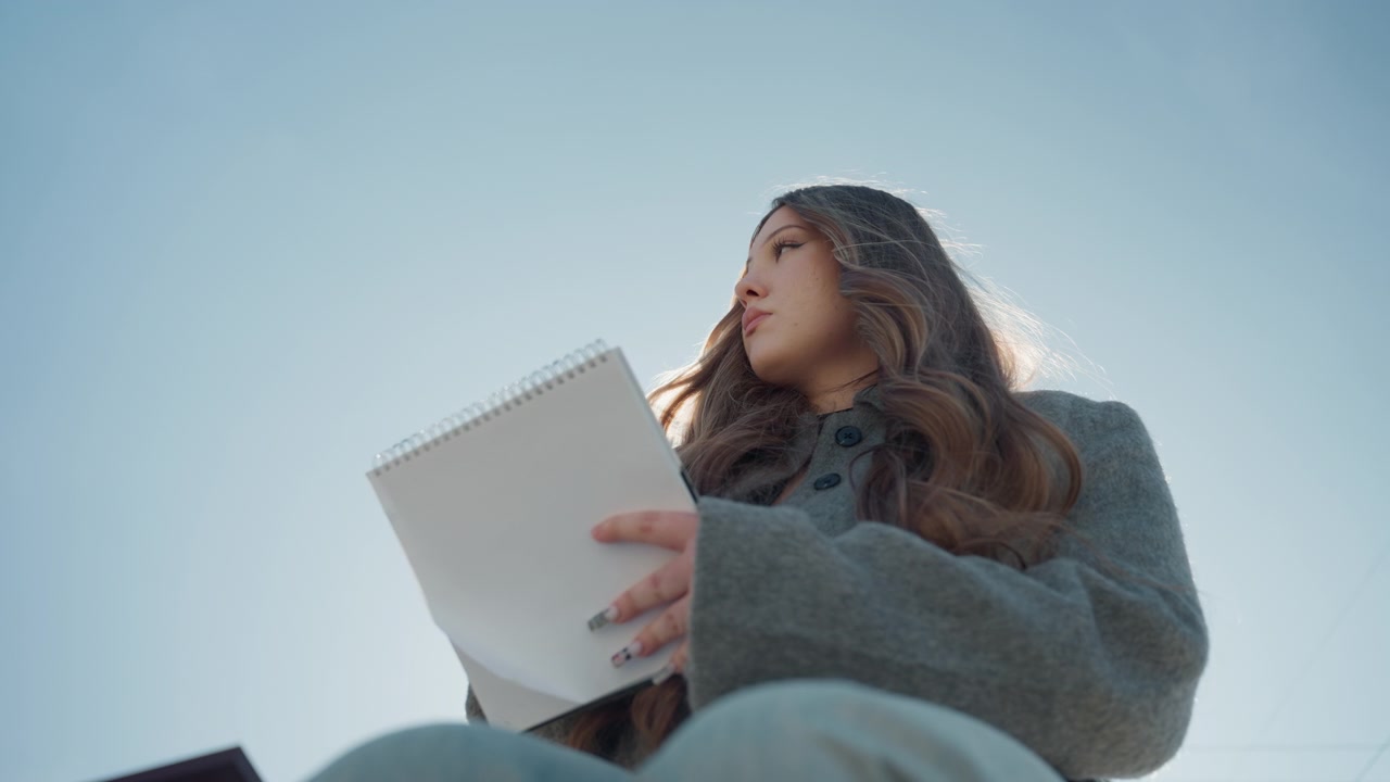 Bella studentessa asiatica che disegna su un diario su una sporgenza all'aperto, retroilluminata dal sole, con cappotto grigio e jeans, espressione calma, ritratto spontaneo dal basso che cattura il bagliore dei capelli e il cielo, un tranquillo momento di studio.