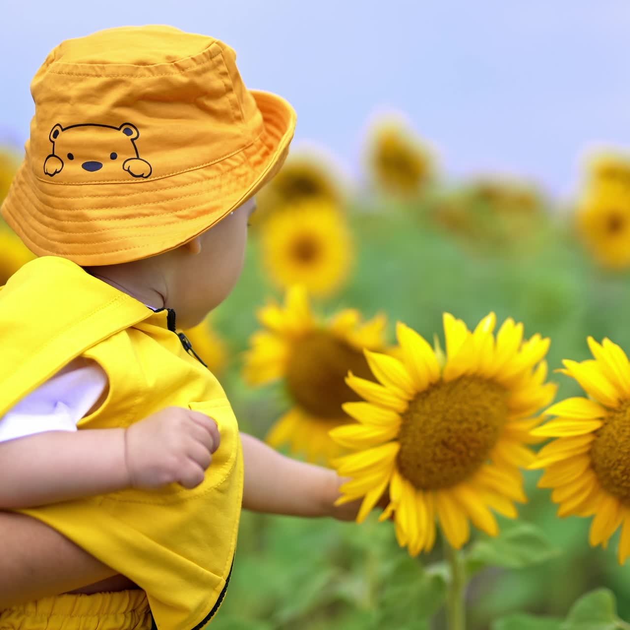 Smiling adorable kid enjoying flowers around him. Mother is holding her son in hands while he touches the sunflowers with interest