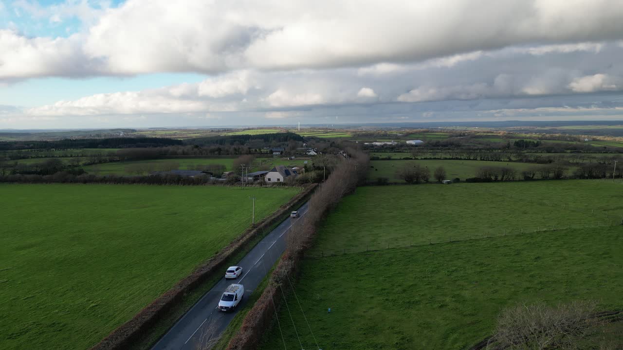 Stationary drone shot capturing a rural road winding through green fields and farmland under a partly cloudy sky. A peaceful British countryside scene.