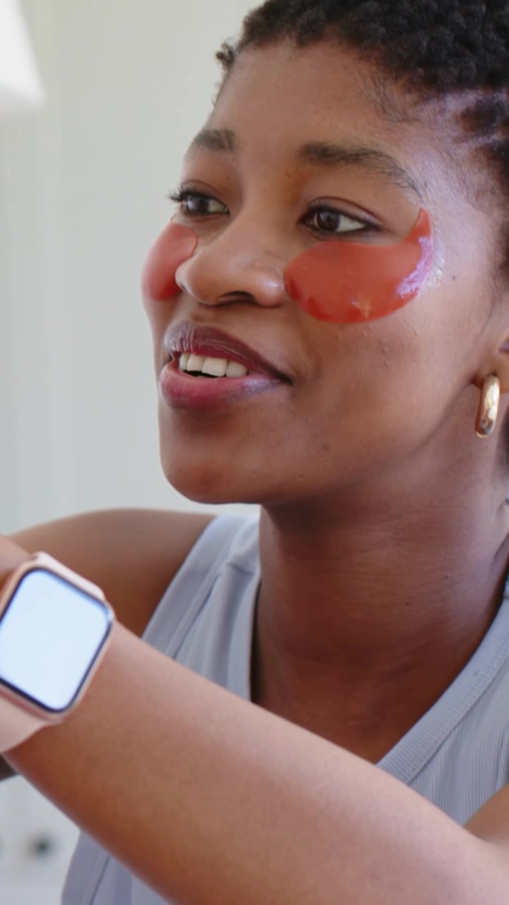 Video verticale: Donna che tira lo specchio, controlla delle patch in gel rosse alla sua toeletta a casa, mostrando un orologio rosa.