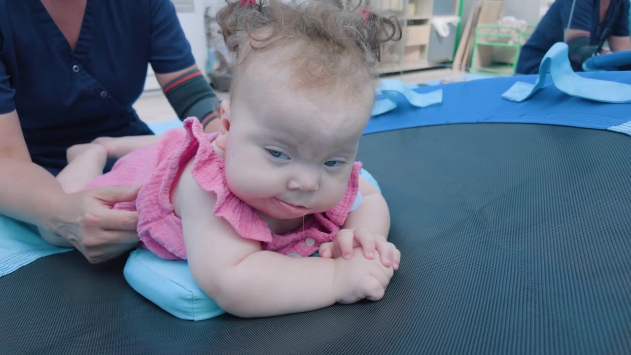 A baby sits with her back to the camera as an adult offers a small toy, another child's leg visible nearby