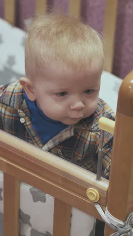 worried little baby leans on edge of wooden cot with soft linen in light children room at home closeup