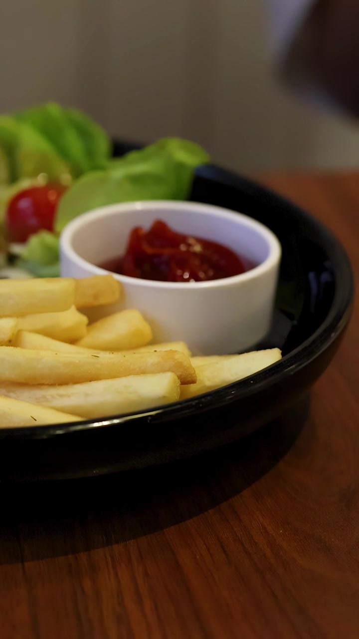 Primo piano di una mano che intinge una patatina fritta nel ketchup accanto a patatine e insalata, inquadratura statica