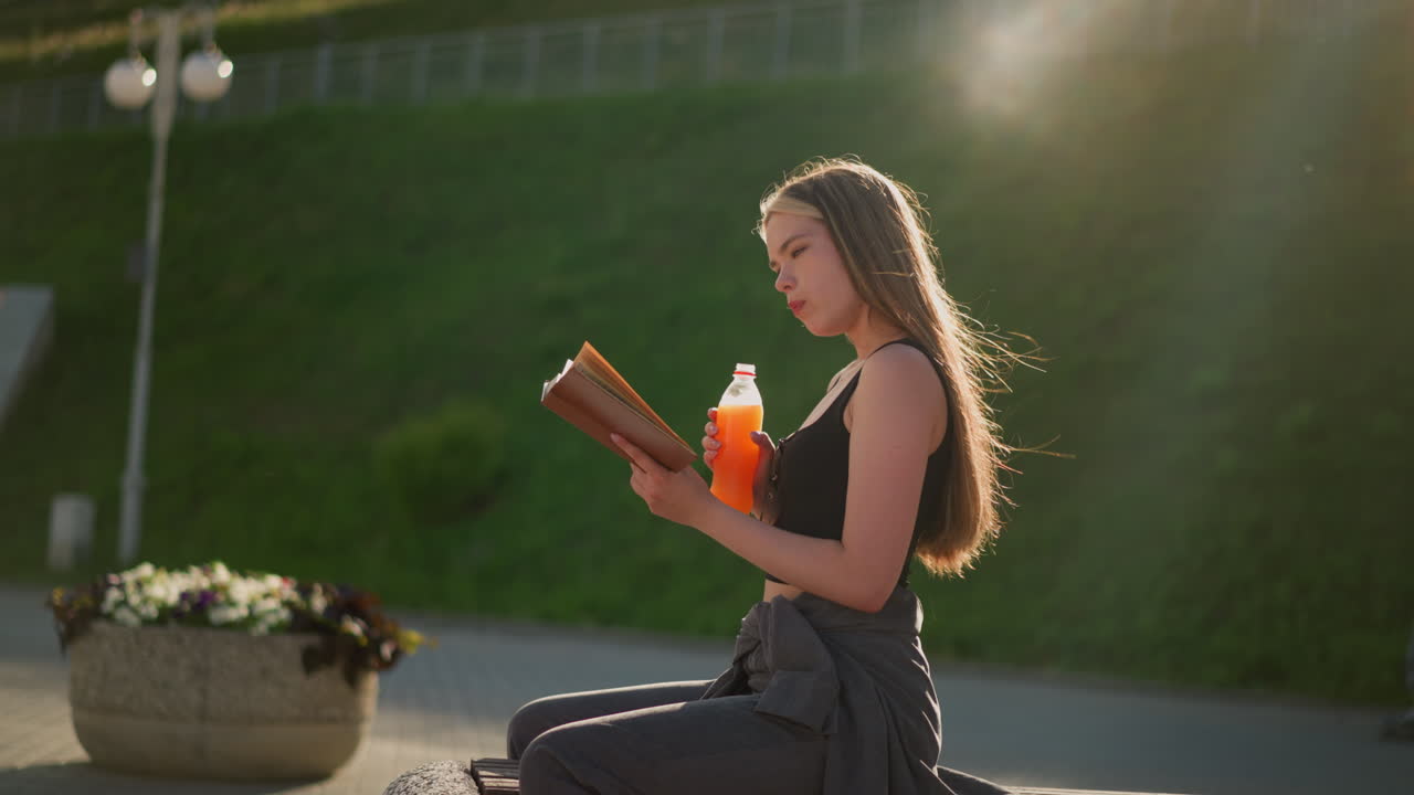 mujer sentada al aire libre, sosteniendo un libro en una mano y bebiendo de una botella de jugo con la otra, continúa leyendo mientras la luz del sol crea un ambiente cálido en el fondo