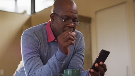 African-american-senior-man-leaning-on-kitchen-counter-using-smartphone,-taking-off-glasses