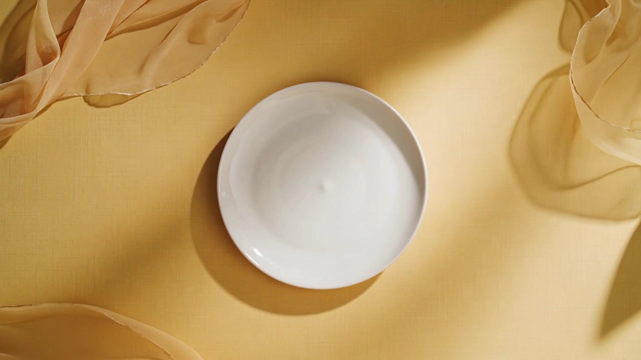 Moving camera pulling white ceramic plate closer on yellow backdrop, showing rim and fabric shadows