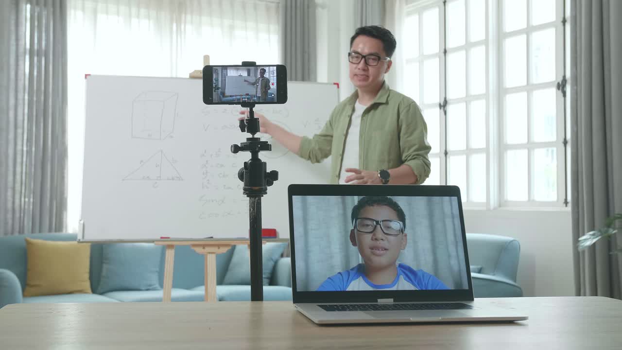 Asian Male Teacher With Glasses Shoots Video By Smartphone Camera While Having Video Call On Laptop And Teaching Math At Home