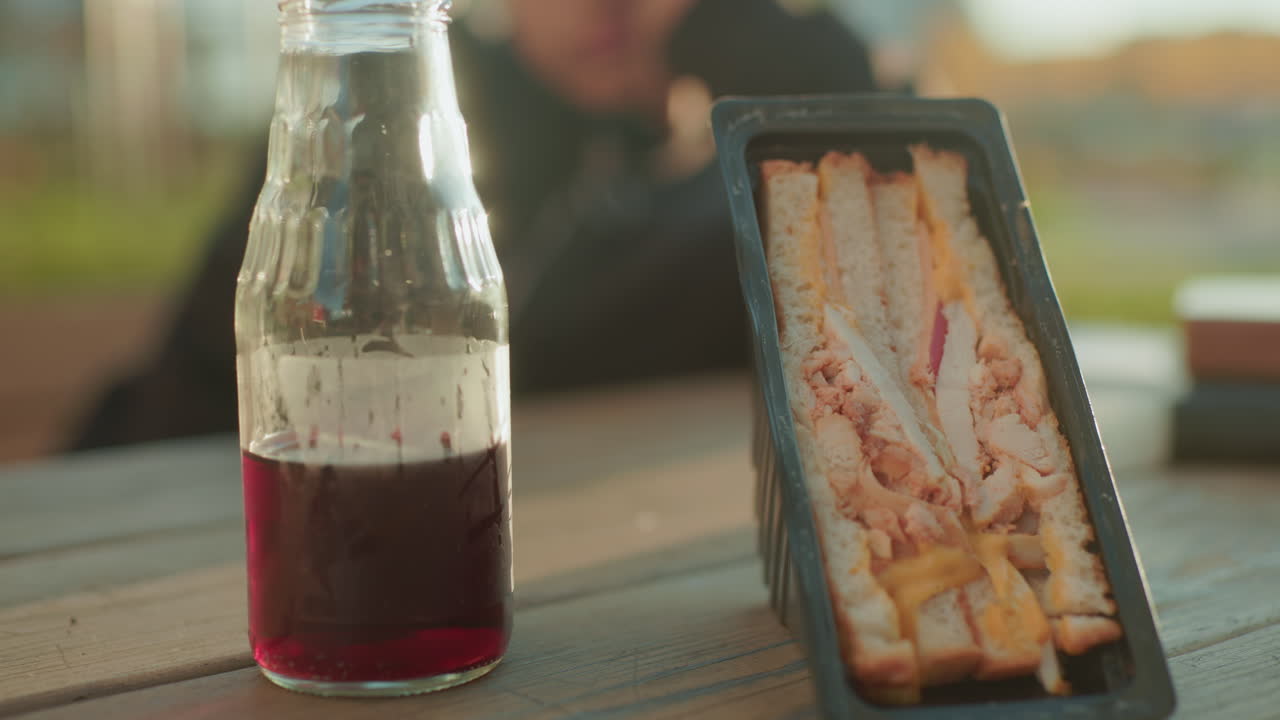 Close up of sandwich pack with layered bread, cheese, and meat placed on wooden table next to glass bottle of juice, warm sunlight casting glow with blurred person in background