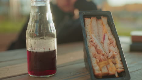 close up of sandwich pack with layered bread, cheese, and meat placed on wooden table next to glass bottle of juice, warm sunlight casting glow with blurred person in background