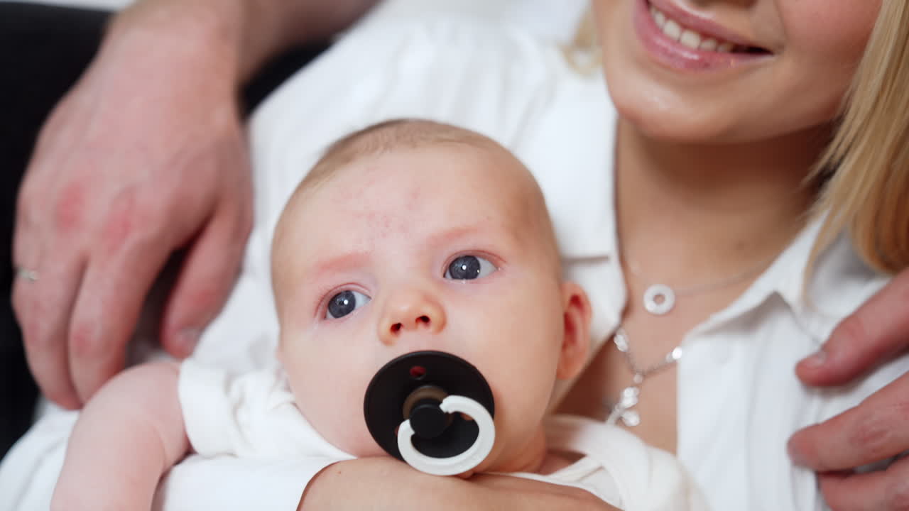 Grey-eyed Caucasian newborn with a black pacifier. Mom waves her son and kisses him on the head. Close up.